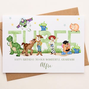 May include: A white birthday greeting card featuring watercolor illustrations of Toy Story characters including Buzz Lightyear, Woody, Jessie, Rex, Bullseye, Mr. Potato Head, and aliens. The word THREE is printed in green with the text Alfie below.