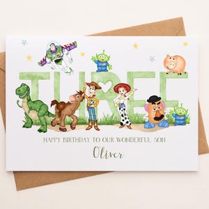 May include: A white birthday card featuring watercolor illustrations of Toy Story characters including Buzz Lightyear, Woody, Jessie, Rex, Bullseye, Mr. Potato Head, and aliens. The word THREE is in large green letters. Text reads: HAPPY BIRTHDAY TO OUR WONDERFUL SON Oliver.