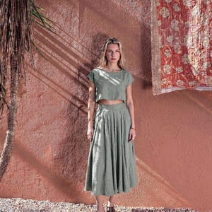 Puede incluir: Una mujer con un top corto de lino verde salvia y una falda midi plisada a juego. Lleva botines marrones. El fondo presenta una pared roja texturizada y un textil decorativo.