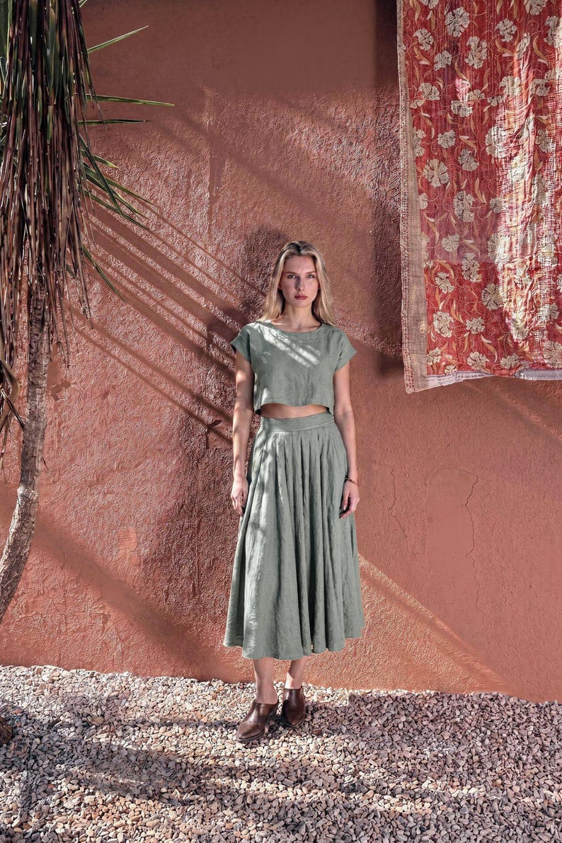 Puede incluir: Una mujer con un top corto de lino verde salvia y una falda midi plisada a juego. Lleva botines marrones. El fondo presenta una pared roja texturizada y un textil decorativo.