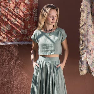 Puede incluir: Conjunto de lino verde salvia de dos piezas. La parte superior es una camisa corta de manga corta. La falda es una falda larga de cintura alta y fluida con bolsillos. Se lleva un collar con un colgante. El fondo presenta una pared texturizada y textiles estampados.