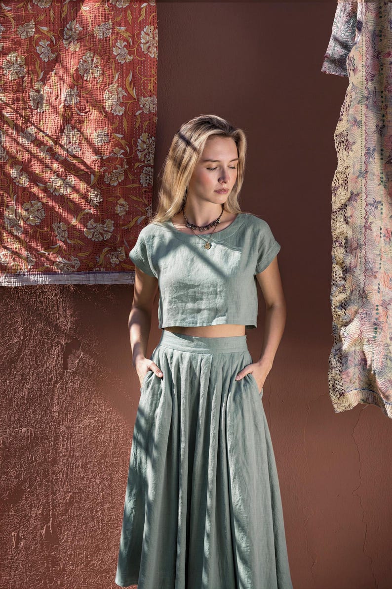 Puede incluir: Conjunto de lino verde salvia de dos piezas. La parte superior es una camisa corta de manga corta. La falda es una falda larga de cintura alta y fluida con bolsillos. Se lleva un collar con un colgante. El fondo presenta una pared texturizada y textiles estampados.