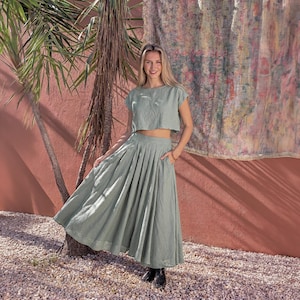 Puede incluir: Conjunto de dos piezas verde salvia compuesto por un top corto y una falda plisada con bolsillos. La tela ligera es ideal para el verano. La modelo est&aacute; de pie frente a una pared roja, con una palmera y un textil estampado en el fondo.