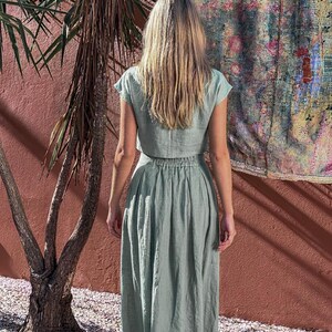 Puede incluir: Un conjunto de dos piezas verde claro, que consta de un top corto y una falda larga. El conjunto est&aacute; hecho de una tela suave y fluida. La modelo est&aacute; de pie frente a una pared roja con un tapiz colorido.
