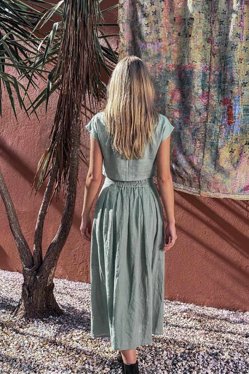 Puede incluir: Un conjunto de dos piezas verde claro, que consta de un top corto y una falda larga. El conjunto est&aacute; hecho de una tela suave y fluida. La modelo est&aacute; de pie frente a una pared roja con un tapiz colorido.