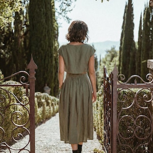 以下が含まれることがあります： オリーブグリーンのクロップドトップスとミディスカートのツーピースを着た人が、カメラから遠ざかっています。黒いブーツを履いています。石畳の小道、装飾的な鉄の門、高い木々がある屋外のシーンです。