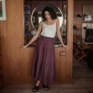 May include: A woman wearing a white camisole top and a tiered, burgundy-colored skirt. The skirt falls to the ankles. She is wearing black sandals. The background is a wooden wall with a round mirror.