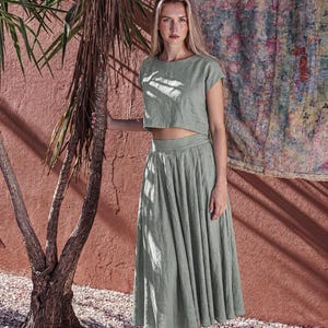 Puede incluir: Un conjunto de dos piezas verde salvia que consta de un top corto y una falda midi. El top tiene mangas cortas y cuello redondo. La falda es plisada y llega a la mitad de la pantorrilla. El conjunto se combina con botines negros. El fondo presenta una pared roja y una palmera.