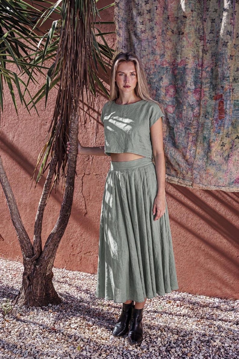 Puede incluir: Un conjunto de dos piezas verde salvia que consta de un top corto y una falda midi. El top tiene mangas cortas y cuello redondo. La falda es plisada y llega a la mitad de la pantorrilla. El conjunto se combina con botines negros. El fondo presenta una pared roja y una palmera.