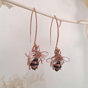 Puede incluir: Pendientes de alambre de cobre con diseños de abeja. Las abejas tienen cuerpos negros y marrones, alas transparentes y cuelgan de largos ganchos de cobre curvados. Los pendientes se muestran sobre un fondo suave y neutro.