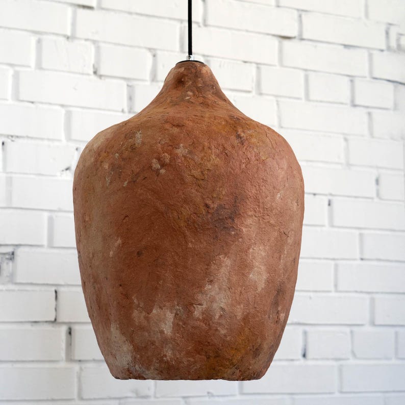 Op de afbeelding: Een hanglamp met een getextureerde, aarde-bruine kap. De kap heeft een afgeronde, bolvormige vorm met een smallere bovenkant, opgehangen aan een zwart snoer tegen een witte bakstenen muur. De lamp is een decoratief element.