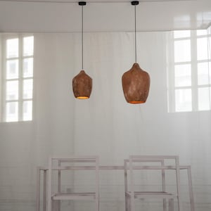 Op de afbeelding: Twee bruine, getextureerde hanglampen hangen boven een tafel en stoelen. De lampen geven een warme gloed. De tafel en stoelen zijn licht en neutraal van kleur.