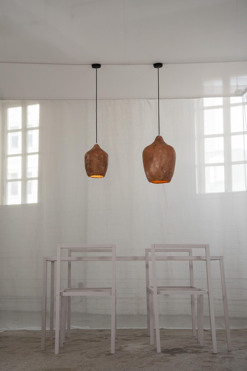 Op de afbeelding: Twee bruine, getextureerde hanglampen hangen boven een tafel en stoelen. De lampen geven een warme gloed. De tafel en stoelen zijn licht en neutraal van kleur.