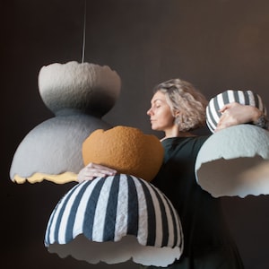 May include: A woman holds up four different handmade paper lampshades. The lampshades are in various colors and patterns, including gray, white, black, and orange.