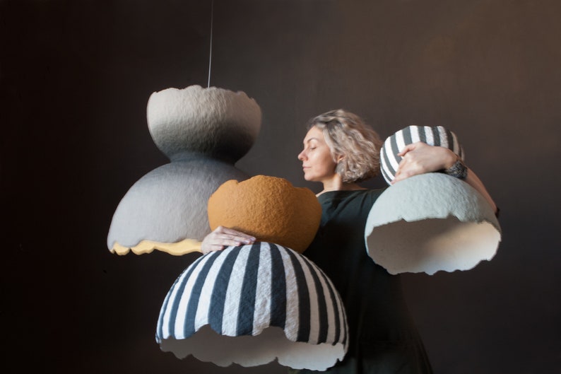 May include: A woman holds up four different handmade paper lampshades. The lampshades are in various colors and patterns, including gray, white, black, and orange.