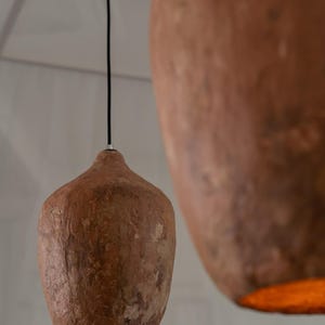 Op de afbeelding: Twee bruine hanglampen met een getextureerde, organische vorm, opgehangen aan zwarte snoeren. De lampen geven aan de onderkant een warme, oranje gloed af. De achtergrond is neutraal wit, wat een minimalistische uitstraling cre&euml;ert.