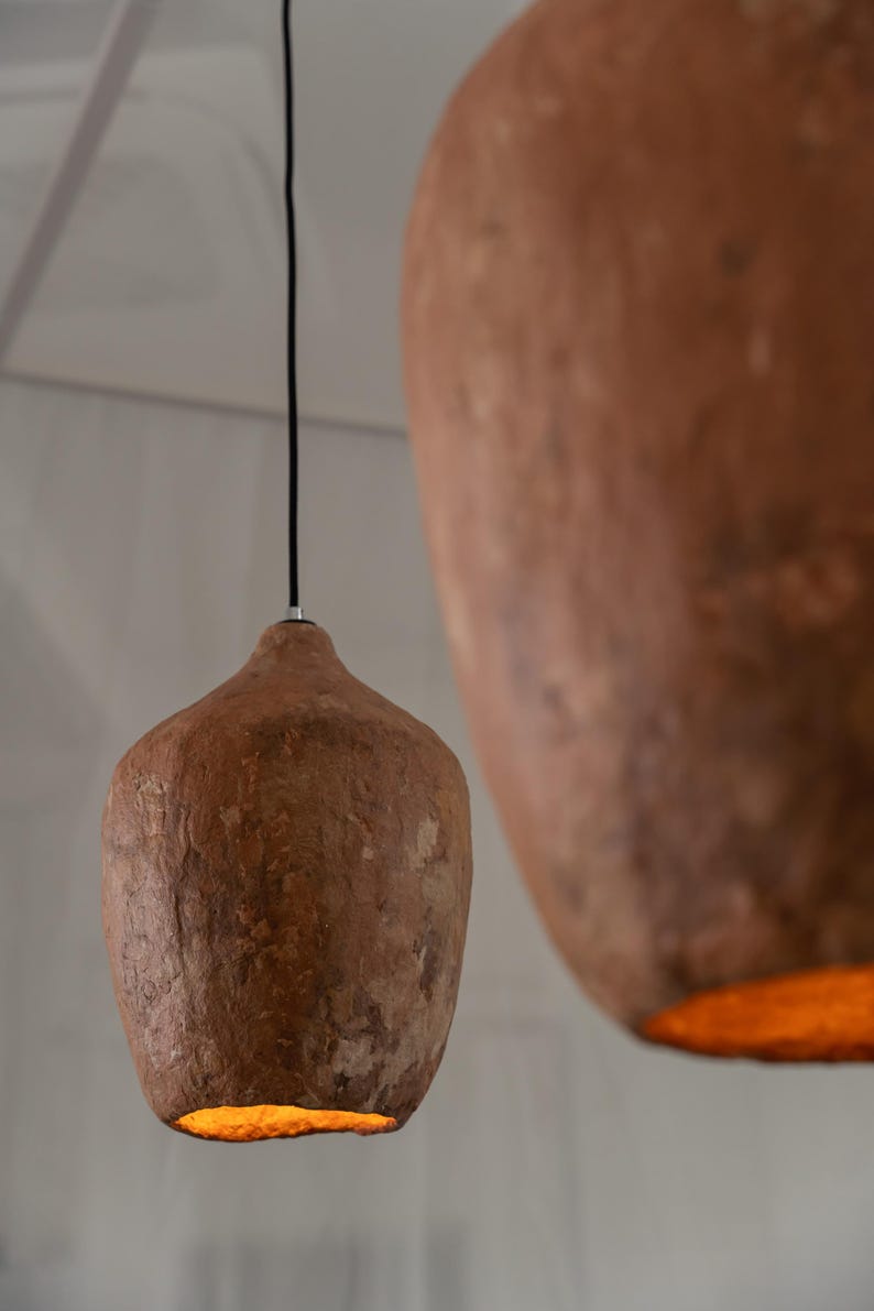 Op de afbeelding: Twee bruine hanglampen met een getextureerde, organische vorm, opgehangen aan zwarte snoeren. De lampen geven aan de onderkant een warme, oranje gloed af. De achtergrond is neutraal wit, wat een minimalistische uitstraling cre&euml;ert.