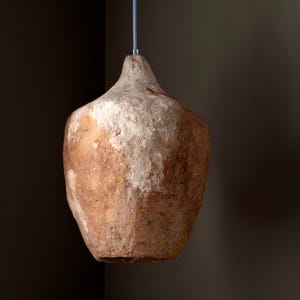 Op de afbeelding: Een hanglamp met een textuur, organische vorm. De lamp heeft een bruin en gebroken witte kleurstelling, met een ruw, ongelijkmatig oppervlak. Hij hangt aan een dun, grijs snoer, wat een rustieke, natuurlijke uitstraling cre&euml;ert.