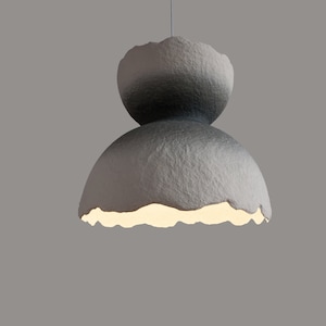 May include: A gray, textured, paper lampshade with a unique, organic shape. The lampshade has a light inside, casting a warm glow.