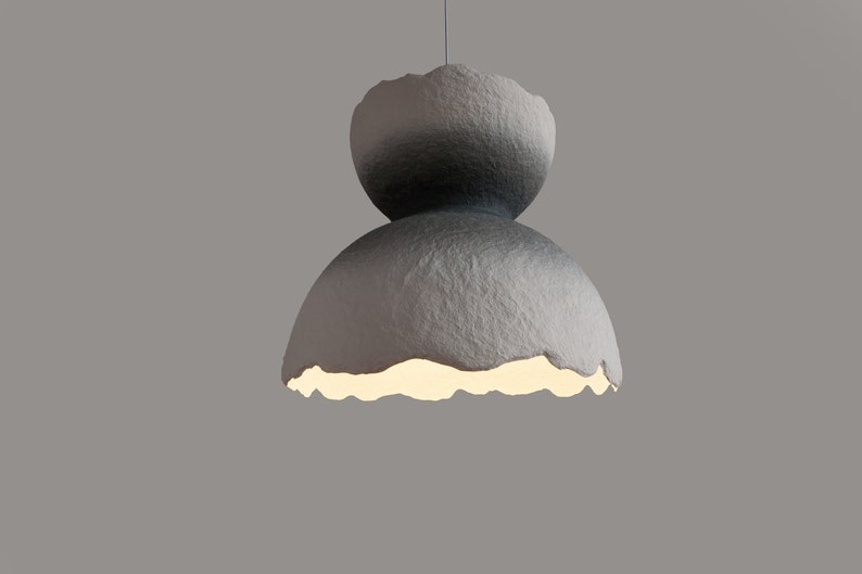 May include: A gray, textured, paper lampshade with a unique, organic shape. The lampshade has a light inside, casting a warm glow.