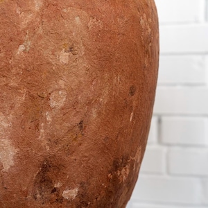 Op de afbeelding: Een groot, getextureerd, roodbruin keramisch vat met een afgeronde vorm. Het oppervlak heeft een ruwe, verouderde uitstraling met variaties in kleur en textuur. De achtergrond is een witte bakstenen muur.