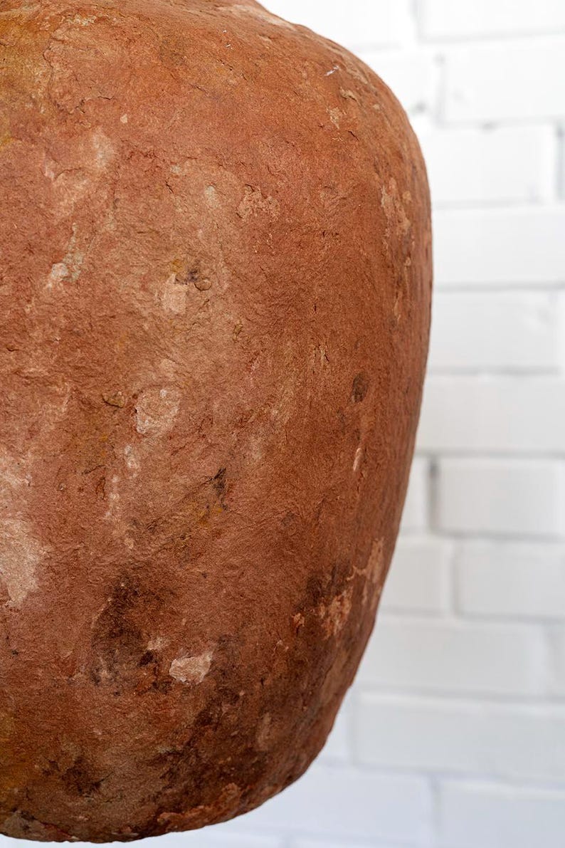 Op de afbeelding: Een groot, getextureerd, roodbruin keramisch vat met een afgeronde vorm. Het oppervlak heeft een ruwe, verouderde uitstraling met variaties in kleur en textuur. De achtergrond is een witte bakstenen muur.