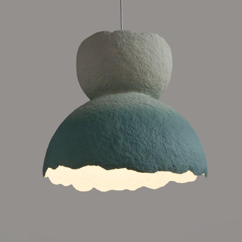 May include: A white and green ombre paper pendant light with a textured surface. The light is turned on and illuminates the room.