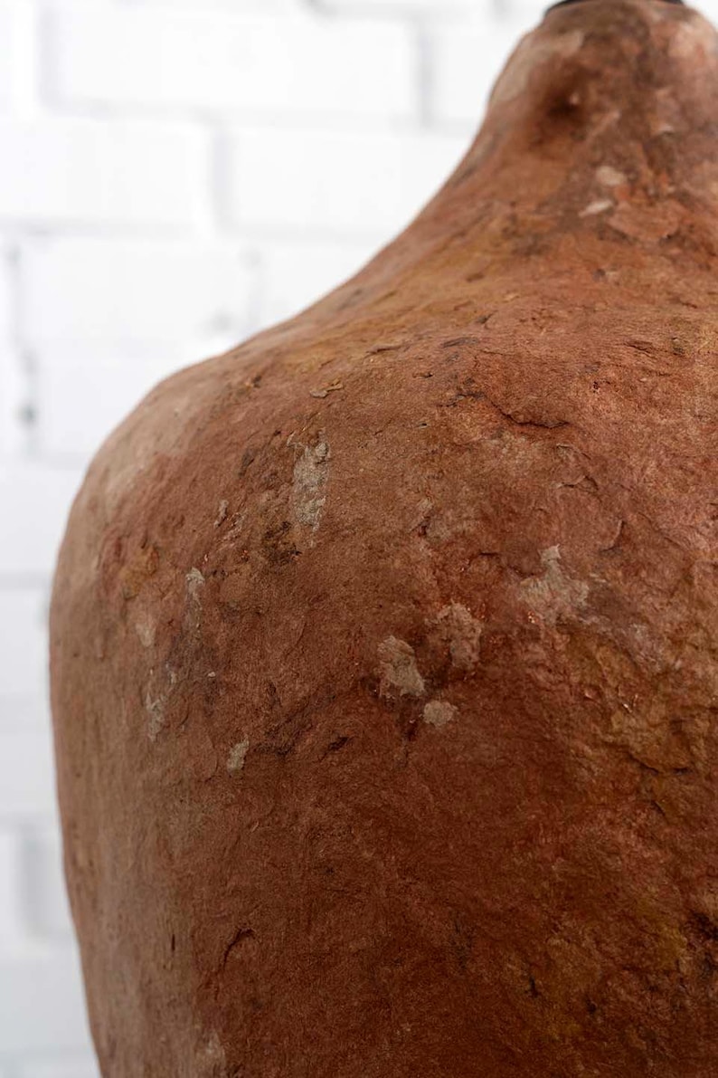Op de afbeelding: Close-up van een grote, getextureerde, roodbruine keramische vaas. Het oppervlak heeft een ruwe, verweerde uitstraling met lichtere vlekken. De vaas heeft een afgeronde schouder en een smalle hals, wat wijst op een rustiek of ambachtelijk ontwerp.