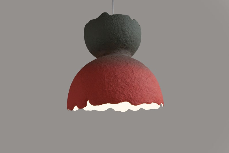 May include: A red and gray paper lampshade with a unique, textured, and slightly irregular shape. The lampshade has a white interior and is hanging from a cord.