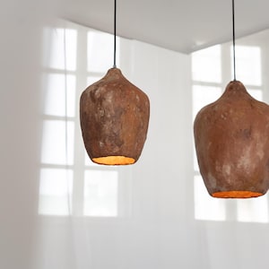 Op de afbeelding: Twee hanglampen met een rustieke, aardse uitstraling. De lampen hebben een druppelvorm en een getextureerd, bruin oppervlak. Het warme licht van de lampen contrasteert met de witte achtergrond.
