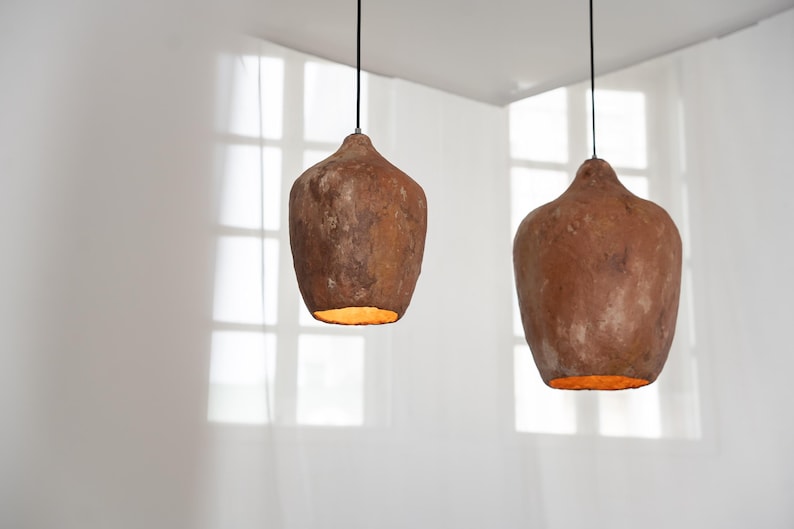 Op de afbeelding: Twee hanglampen met een rustieke, aardse uitstraling. De lampen hebben een druppelvorm en een getextureerd, bruin oppervlak. Het warme licht van de lampen contrasteert met de witte achtergrond.
