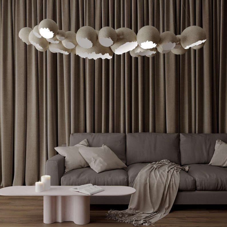 May include: A living room with a brown sofa, two throw pillows, a throw blanket, and a light pink coffee table with two candles on it. The room has brown curtains and a unique light fixture made of many round, beige, paper-like orbs with cutouts.