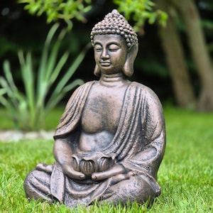 Può includere: Statua di Buddha color bronzo in posizione di meditazione seduta, con un fiore di loto. La statua è su un prato erboso, con uno sfondo verde sfocato di alberi e fogliame. La statua ha un'espressione serena.