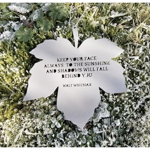 Puede incluir: Un adorno de hoja de metal plateado con la cita "Mantén siempre tu rostro hacia el sol y las sombras caerán detrás de ti" de Walt Whitman.