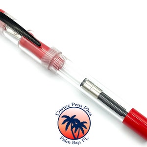 May include: A clear fountain pen with red accents and a black clip. The pen has a transparent body, allowing a view of the internal components. The pen's design includes a red cap and end, and the brand logo is visible.