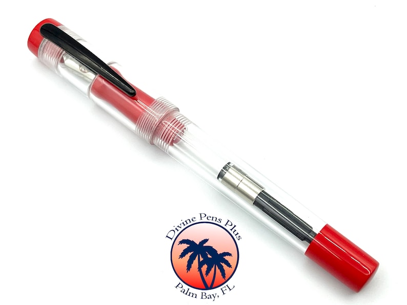 May include: A clear fountain pen with red accents and a black clip. The pen has a transparent body, allowing a view of the internal components. The pen's design includes a red cap and end, and the brand logo is visible.
