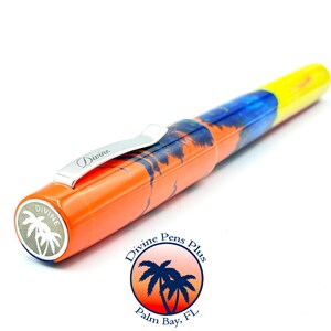 May include: A colorful fountain pen with a tropical theme. The pen features an orange barrel with a palm tree design, a blue and yellow body, and a silver clip. The pen has the word "Divine" on the clip and the brand logo on the end.