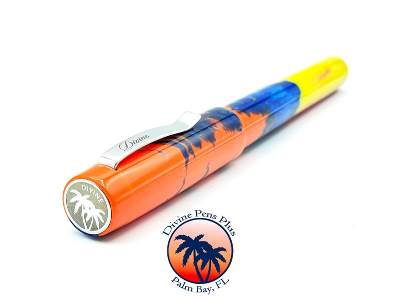 May include: A colorful fountain pen with a tropical theme. The pen features an orange barrel with a palm tree design, a blue and yellow body, and a silver clip. The pen has the word "Divine" on the clip and the brand logo on the end.