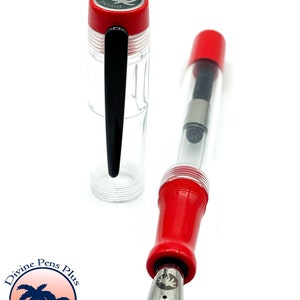 May include: A clear fountain pen with red accents and a silver nib. The pen's cap is red with a silver emblem and the text "DIVINE". The pen is displayed on a white background. The pen is from Divine Pens Plus, Palm Bay, FL.