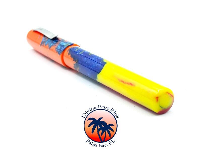 May include: A colorful pen with an orange cap, a blue and orange patterned section, and a yellow barrel. The pen has a silver clip and is set against a white background. The pen is from Divine Pens Plus, Palm Bay, FL.