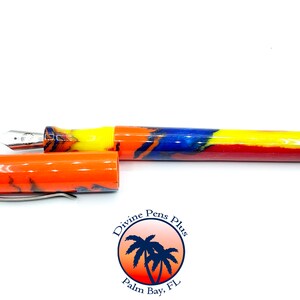 May include: A colorful fountain pen with an open cap. The pen features a silver nib and a vibrant barrel design with orange, yellow, blue, and red hues. The pen's cap is orange with a silver clip. The logo "Divine Pens Plus" is visible.