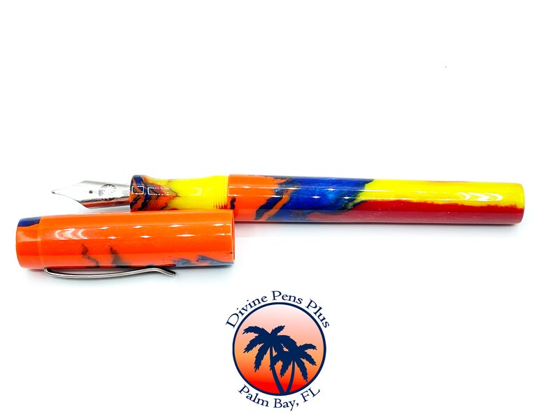 May include: A colorful fountain pen with an open cap. The pen features a silver nib and a vibrant barrel design with orange, yellow, blue, and red hues. The pen's cap is orange with a silver clip. The logo "Divine Pens Plus" is visible.