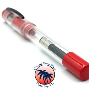 May include: A clear fountain pen with red accents, a black clip, and a red cap. The pen's internal components are visible through the transparent barrel. The pen has a black nib and a silver-colored metal piece. The pen is labeled "Divine Pens Plus, Palm Bay, FL."
