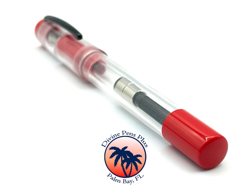 May include: A clear fountain pen with red accents, a black clip, and a red cap. The pen's internal components are visible through the transparent barrel. The pen has a black nib and a silver-colored metal piece. The pen is labeled "Divine Pens Plus, Palm Bay, FL."