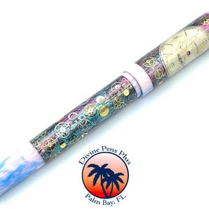 Puede incluir: Un bol&iacute;grafo decorativo con un dise&ntilde;o steampunk. El bol&iacute;grafo presenta una esfera de reloj y engranajes sobre un fondo turquesa, morado y dorado. La punta del bol&iacute;grafo es azul y blanca, y el bol&iacute;grafo tiene una banda blanca. El logotipo de Divine Pens Plus es visible.
