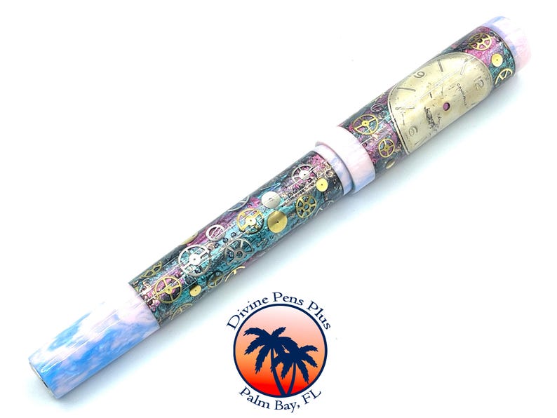 Puede incluir: Un bol&iacute;grafo decorativo con un dise&ntilde;o steampunk. El bol&iacute;grafo presenta una esfera de reloj y engranajes sobre un fondo turquesa, morado y dorado. La punta del bol&iacute;grafo es azul y blanca, y el bol&iacute;grafo tiene una banda blanca. El logotipo de Divine Pens Plus es visible.