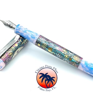 Puede incluir: Una pluma estilogr&aacute;fica decorativa con plum&iacute;n plateado y cuerpo adornado. El cuerpo presenta un dise&ntilde;o de engranajes y elementos mec&aacute;nicos en tonos dorados, turquesa y morado. La tapa de la pluma est&aacute; abierta, mostrando el plum&iacute;n y la palabra "Divine".