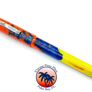 May include: A colorful fountain pen with an orange cap and a silver clip. The barrel features a mix of blue, orange, and yellow hues. The pen has the text "Divine Pens Plus" and a logo with palm trees.