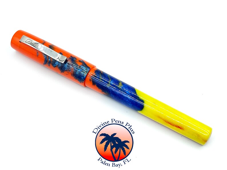 May include: A colorful fountain pen with an orange cap and a silver clip. The barrel features a mix of blue, orange, and yellow hues. The pen has the text "Divine Pens Plus" and a logo with palm trees.