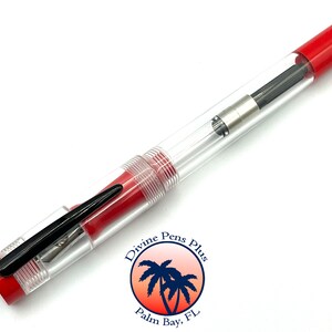 May include: A clear fountain pen with red accents. The pen has a transparent barrel, a black nib, and a red cap and end. The pen has a clip and a silver-colored metal section. The pen is a writing instrument.